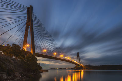 Illuminated bridge over river against cloudy sky at dusk