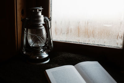 Cozy atmosphere of room with retro oil lantern and opened book placed on windowsill with misted window in house