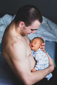 Shirtless father holding son while sitting at home