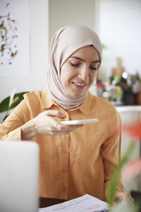 Smiling woman with hijab using cell phone and scanning bills