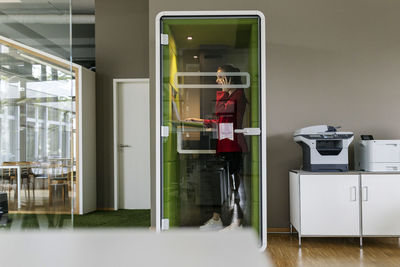 Female entrepreneur talking on smart phone while standing in soundproof cabin at office