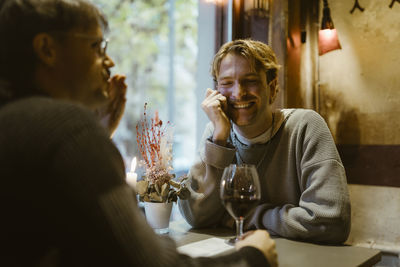 Smiling young gay man talking with boyfriend during candlelight date at restaurant