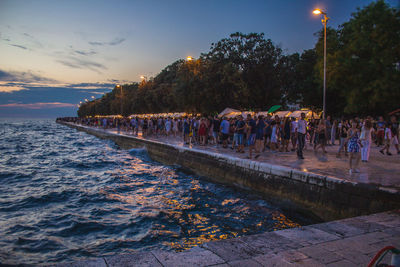 People on street by illuminated trees against sky at sunset