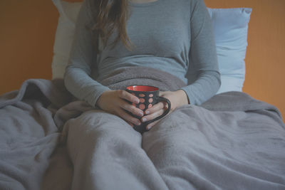 Midsection of woman holding coffee cup while sitting on bed at home