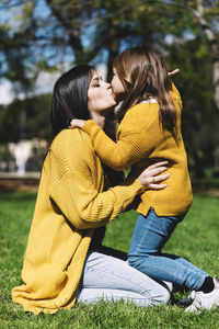 Side view of mother kissing daughter while kneeling on grassy field in park