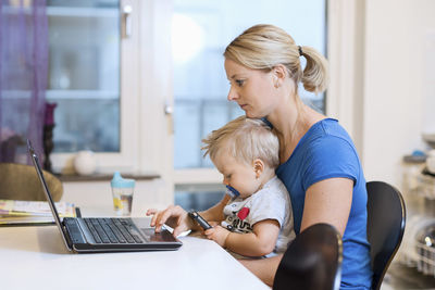 Mother working on laptop while baby boy using smart phone on her lap at home