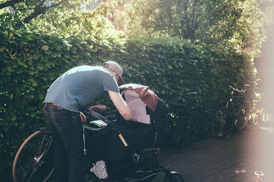 Full length of father bending over baby carriage on footpath at park during sunny day