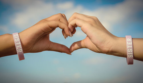 Cropped hands of sisters making heart shape against sky