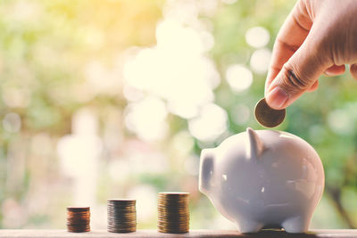 Cropped hand putting money into piggy bank by coins stack on table