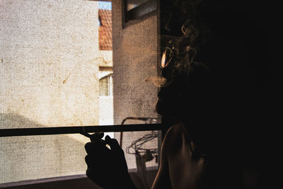 Silhouette young man smoking cigarette while sitting by window at home