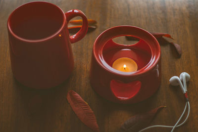 Red cup on wooden table with burning candle, scattered leaves and headphones
