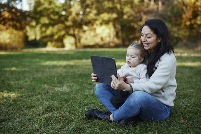 Mother and daughter sitting in autumn park and using digital tablet