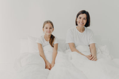 Portrait of smiling mother and daughter sitting on bed against wall at home