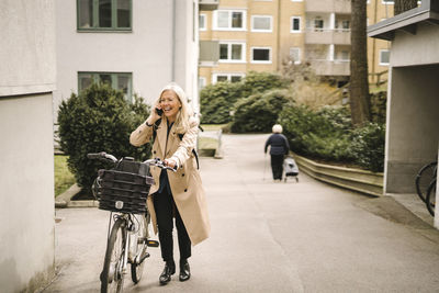 Happy businesswoman talking on smart phone while wheeling bicycle on footpath