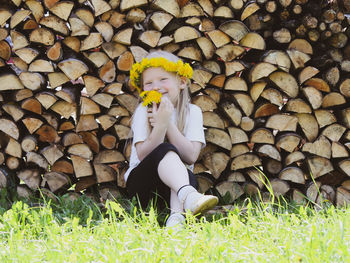 A little girl with a wreath on her head sitting near the firewood stack outdoors.