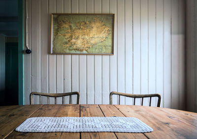 In an old-fashioned room, two old chairs stand at a wooden table on which lies a crocheted doily