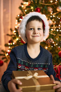 A charming boy in a santa claus hat sits next to the christmas tree with a gift box in his hands. 