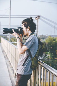 Young man photographing on footbridge against sky