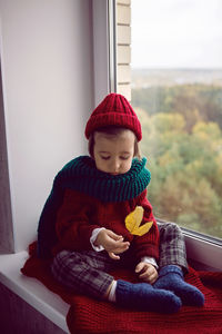 Boy child in red knitted sweater and hat sitting on the window in autumn