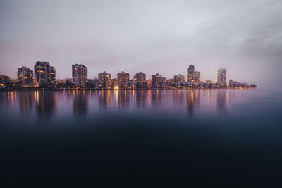 Illuminated buildings by river against sky in city