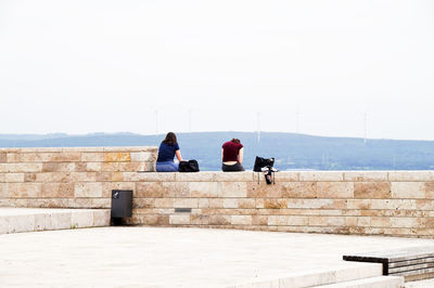 Rear view of people sitting on wall against sky