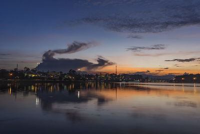 Illuminated factory by lake against sky during sunset