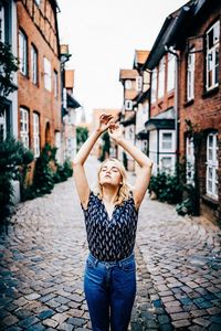 Young woman with eyes closed and arms raised standing on footpath amidst buildings
