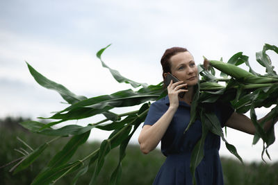 Woman holding leaf while using phone against sky