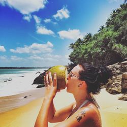 Side view of woman drinking coconut water at beach against sky