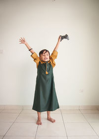 Boy holding game controller while standing with arms outstretched against wall