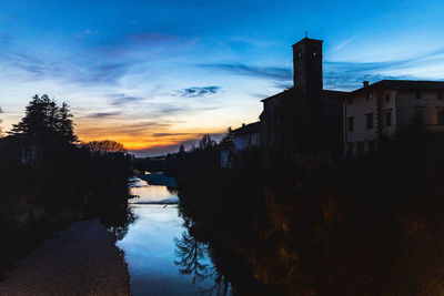 Silhouette buildings by canal against sky at sunset