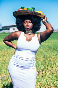 Stylish plus size african woman in white dress with curly hair holding hand on waist and looking at camera while carrying metal plate with bell pepper and tomatoes on head on blurred background of grassy field and blue sky