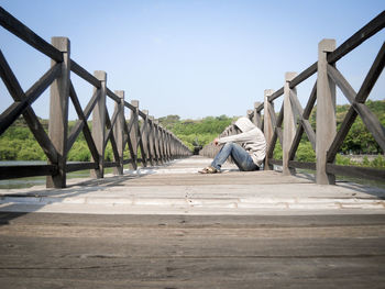 Man in hood sitting on footbridge against clear sky