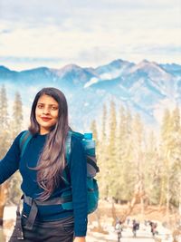 Portrait of smiling young woman standing against mountains and sky during winter