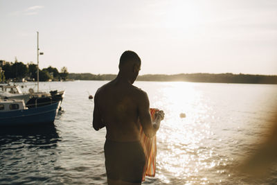 Rear view of shirtless wet man with towel at lake in summer