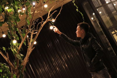 Young man touching electric bulb hanging from tree