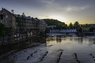 Bridge over river by buildings against sky at sunset