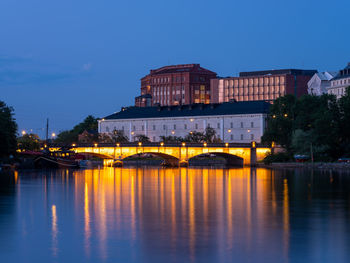 Illuminated buildings by river against blue sky at dusk