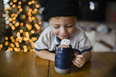Happy boy looking in cup with marshmallow snowman at home