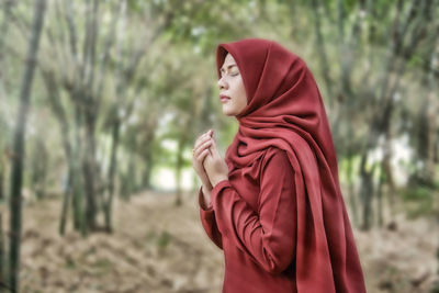 Side view of mid adult woman in hijab praying while standing against trees