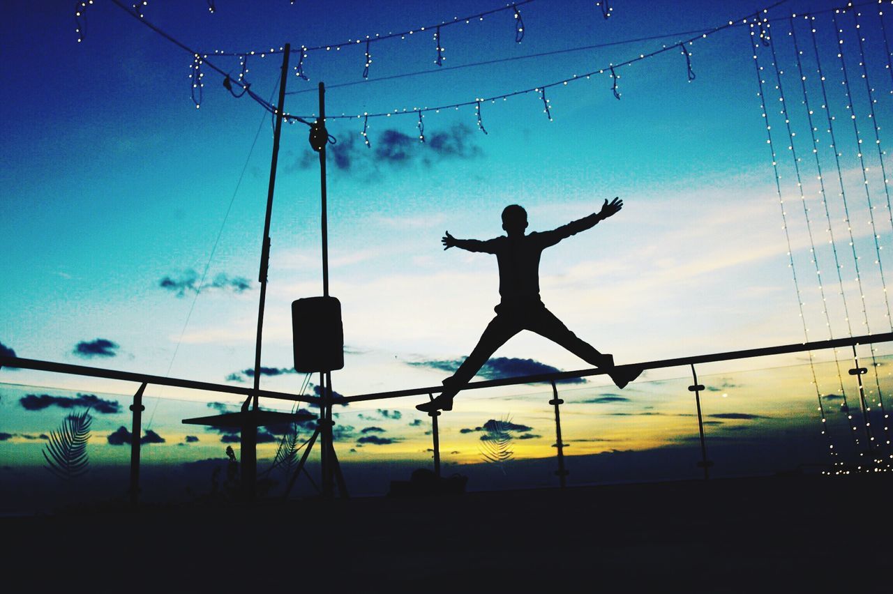 silhouette, sunset, leisure activity, motion, lifestyles, full length, scenics, blue, enjoyment, fence, fun, tranquil scene, sky, mid-air, outdoors, nature, carefree, chain link fence, beauty in nature, outline, cloud - sky, focus on foreground, tranquility