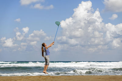 Girl holding butterfly net while standing at beach against sky