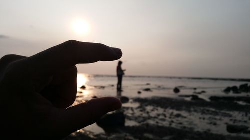 Optical illusion of hand holding person standing at beach against sky during sunset