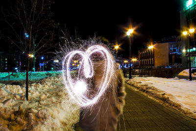 Man making heart shape with illuminated sparklers on street in city at night