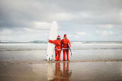Rear view of couple wearing santa costumes holding surfboards in sea