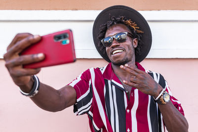 Funky young african american guy in stylish outfit and accessories standing near colorful wall and taking selfie on smartphone on urban street