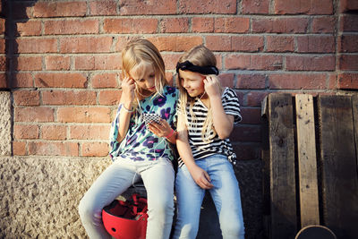 Smiling friends listening music while sitting against brick wall