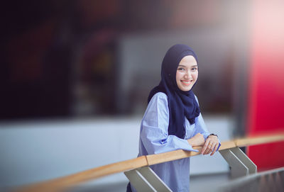 Portrait of teenage girl in hijab standing by railing in corridor