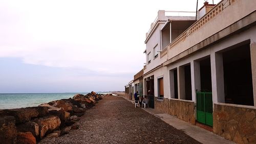 Footpath amidst buildings by sea against sky