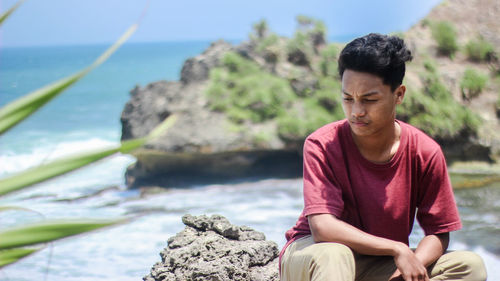 Young man looking away while sitting on rock by sea